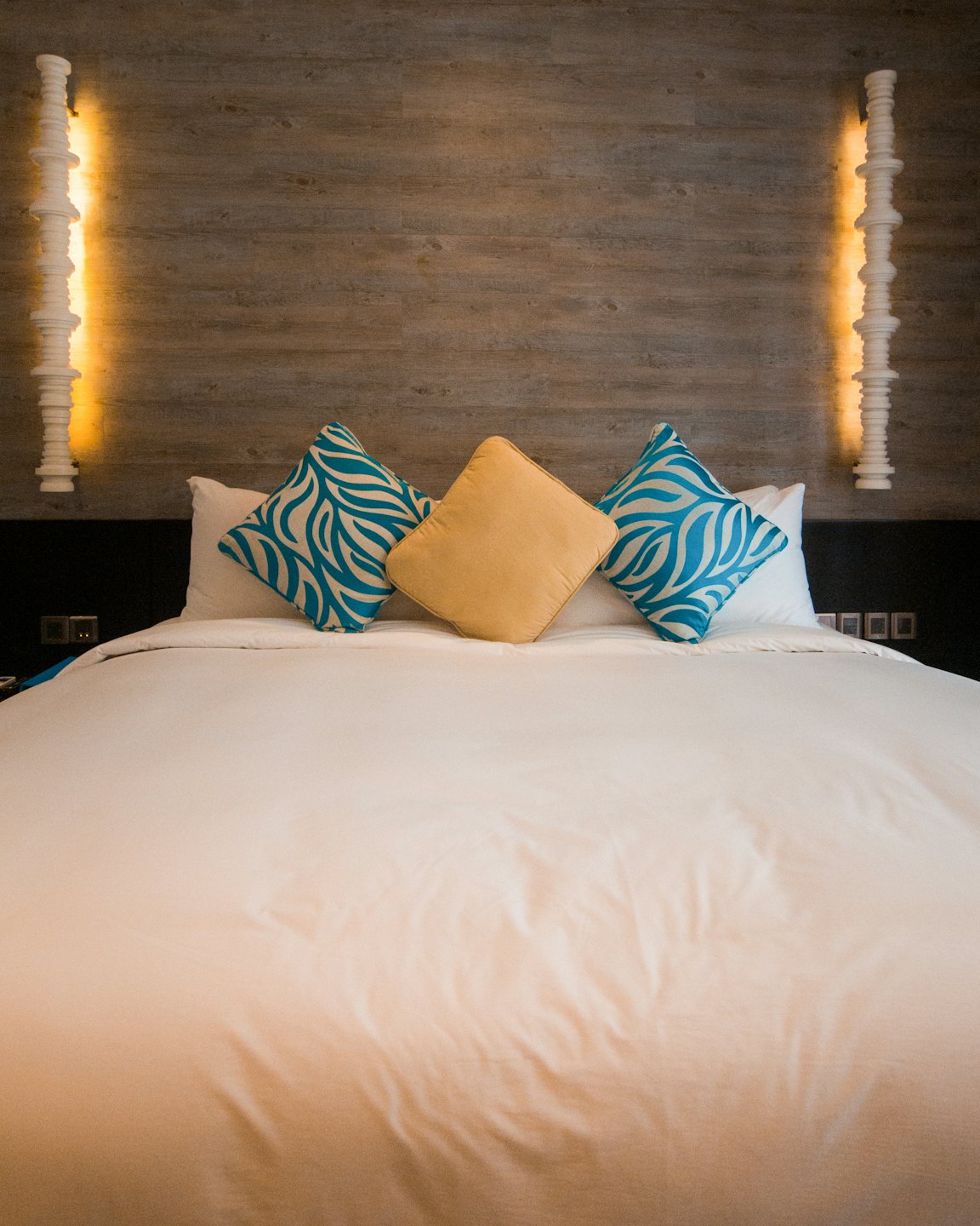 Ein gemachtes Bett mit blauen und gelben Kissen vor einer Holzwand.